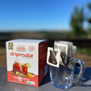 May include: A box of mate tea bags with the brand name "dripmate max" and the text "mate tea bag orgánico" on the front. The box is sitting on a wooden surface next to a clear glass mug with a tea bag inside.