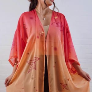 Puede incluir: Un kimono ligero de frente abierto con un degradado que va desde un rosa coral intenso en la parte superior hasta un tono melocotón suave en el dobladillo. La tela está adornada con un estampado celestial de estrellas, constelaciones y soles.