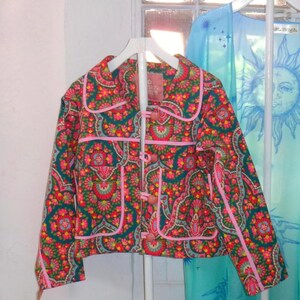Puede incluir: Una chaqueta de manga larga con base verde azulado oscuro y un patrón floral y geométrico repetitivo en tonos rosa, naranja y verde. La chaqueta incluye un cuello redondeado, ribetes de color rosa y cuatro botones redondos rosas al frente.