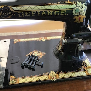 May include: A black vintage sewing machine with gold and green accents. The machine is labeled "DEFIANCE." The machine is sitting on a wooden table.