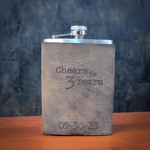 Peut inclure: Une flasque métallique rectangulaire recouverte d'un matériau gris mat aspect cuir. L'avant est gravé avec le texte Cheers to 3 years et la date 09-30-23. Elle possède un bouchon à vis argenté avec un bras articulé sur le dessus.