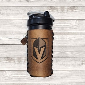 May include: A black and gray water bottle with a leather sleeve featuring the Vegas Golden Knights logo. The brown leather sleeve is stitched and has a small tag attached. The bottle has a black lid with a gray spout.