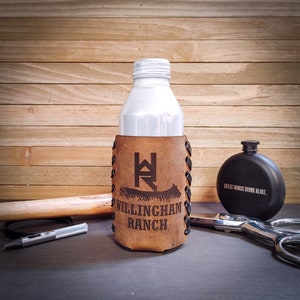 May include: A brown leather drink holder with the text "WILLINGHAM RANCH" and a logo, holding a white bottle. A black flask with the text "GREAT MINDS DRINK ALIKE" is to the right. Other items include scissors and a metal tool.