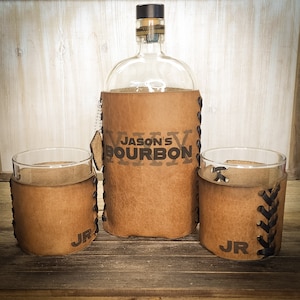 Pode incluir: Uma garrafa de bourbon de vidro transparente e dois copos a condizer, cada um envolto em couro castanho. A garrafa e os copos são marcados com "JASON'S BOURBON" e "JR". O couro é costurado com fio preto, criando uma estética rústica.