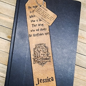Könnte beinhalten: Ein personalisierter Leder-Lesezeichen mit dem eingravierten Namen "Jessica". Das Lesezeichen zeigt das Gryffindor-Wappen und ein Zitat. Oben ist eine gelb-schwarze Quaste befestigt. Das Lesezeichen liegt auf einem dunkelblauen Buch.