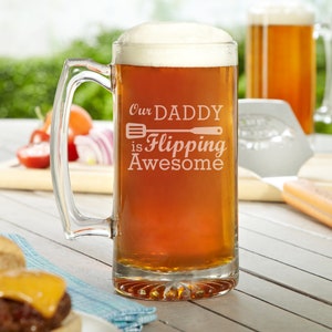 May include: A clear glass beer stein with a handle, filled with a dark amber beer. The stein is engraved with the text "Our Daddy is Flipping Awesome" and a spatula graphic.