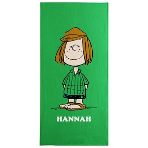 May include: A green beach towel featuring a cartoon illustration of a girl with brown hair, a brown hat, and a green striped shirt. The name "HANNAH" is printed in white at the bottom of the towel.
