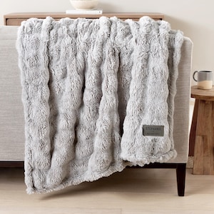 May include: A grey faux fur throw blanket with a textured, plush appearance, draped over a grey sofa. The blanket has a tag that reads "Stephanie" and "Best New Life".
