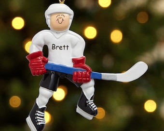 May include: A hockey player ornament with a white jersey and the name "Brett". The figure wears a white helmet, red gloves, black shorts, and black skates. The hockey stick is blue and white. A gold string is attached for hanging.
