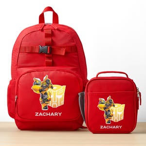 May include: A bright red backpack and matching lunch bag set featuring a yellow and black Bumblebee character graphic, a yellow Autobot logo, and the name ZACHARY printed in white. Both items have zippers and a structured design.