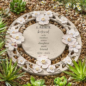 May include: A round, grey stone garden stepping stone with a white floral design. The stone has the text "Laurie beloved wife mother sister daughter aunt friend 1949 - 2024" inscribed on it.