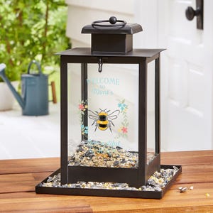 May include: Black metal bird feeder with a rectangular base and a square top. The feeder has glass sides and a decorative panel with a bee illustration and the text "WELCOME to Mimi's". The feeder is filled with birdseed.
