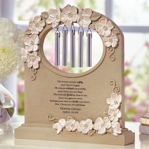 May include: A beige memorial wind chime with a floral design. The chime has a central opening with five silver chimes. The text reads "The breeze sweeps softly...Victoria Carson 1962-2026."
