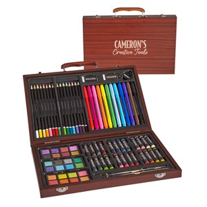 May include: A wooden art case with a handle, labeled "CAMERON'S Creative Tools". The case is open, revealing a variety of art supplies, including colored pencils, markers, crayons, watercolor paints, and a paintbrush.