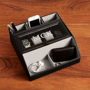 May include: Black leather valet tray with a gray felt interior. The tray has compartments for a phone, watch, wallet, and other accessories. The tray is personalized with the initials "MCK".