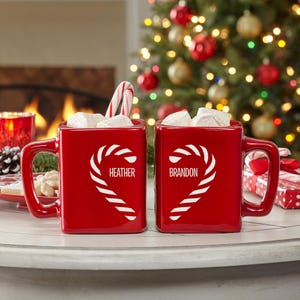 May include: Two red square mugs filled with marshmallows and a candy cane. Each mug has a white candy cane heart design with a name, "Heather" and "Brandon". The mugs are on a white surface with a Christmas tree in the background.