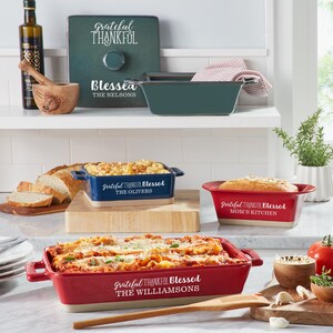 May include: Three ceramic baking dishes with handles in red, blue and green. Each dish has a white inscription that reads "Grateful Thankful Blessed" followed by a family name. The dishes are on a wooden worktop.
