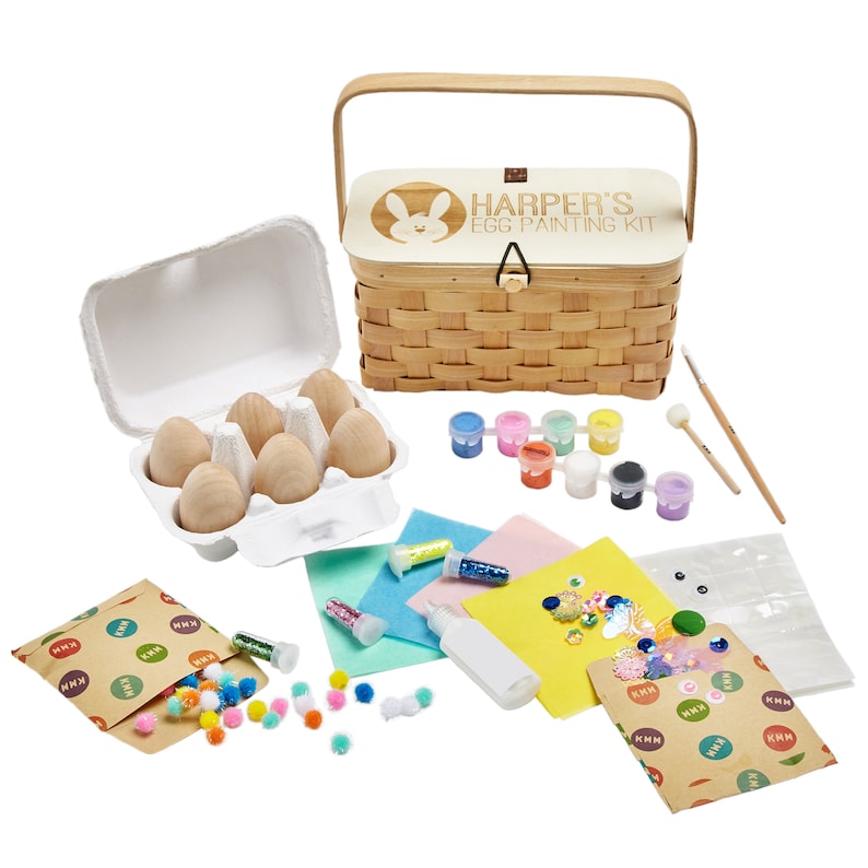 Puede incluir: Una cesta de madera con asa, etiquetada como "Harper's Egg Painting Kit" con un dise&ntilde;o de conejo. La cesta contiene huevos de madera, pintura, purpurina, pompones y otros materiales de manualidades.