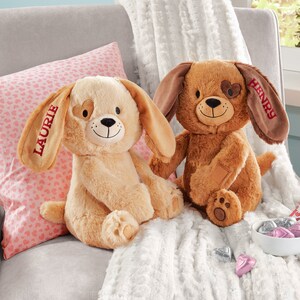 May include: Two plush brown and beige dog toys with embroidered hearts on their eyes. The dog on the left has the name "Laurie" embroidered on its ear and the dog on the right has the name "Henry" embroidered on its ear.