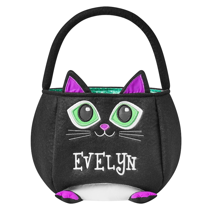May include: Black felt Halloween trick-or-treat bag shaped like a cat. The bag has a black handle and features a cat face with green eyes, purple ears, and the name "EVELYN" embroidered in white. The interior has a green and white polka dot pattern.