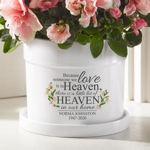 Peut inclure: Pot de fleurs en céramique blanche avec soucoupe, rempli de plantes à fleurs roses et vertes. Le pot porte l'inscription "Because someone we love is in Heaven, there is a little bit of HEAVEN in our home." et "NORMA JOHNSTON 1947-2026."