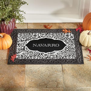 May include: A black and white doormat with the name "NAVARRO" in white lettering. The mat has a decorative border and a central design with swirling patterns and skull accents. Pumpkins and fall foliage surround the mat.