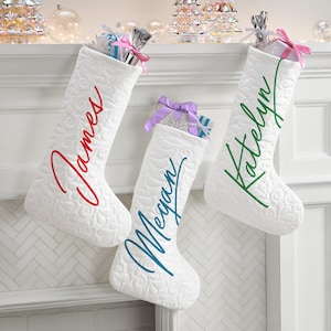 May include: Three white Christmas stockings with different names embroidered on them. The names are James, Megan, and Katelyn. The stockings are hanging on a white mantel.
