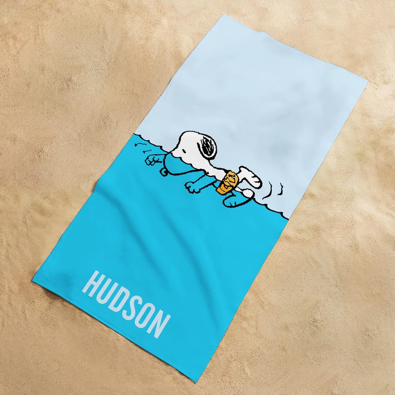 May include: A rectangular beach towel with a light blue and white design. The towel features a cartoon illustration of Snoopy swimming in the ocean. The word "HUDSON" is printed in white on the lower portion of the towel.