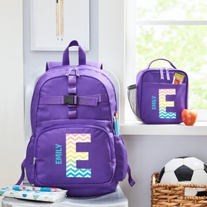 May include: Purple backpack with a rainbow chevron letter E and the name Emily on it. A matching purple lunch bag with a rainbow chevron letter E and the name Emily on it.