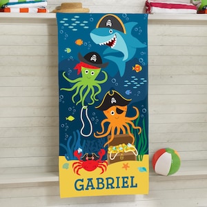 May include: A blue beach towel featuring a cartoon pirate theme. The design includes a shark, octopus, and crab, all wearing pirate hats, along with a treasure chest and the name "GABRIEL". A colorful beach ball is on the right.