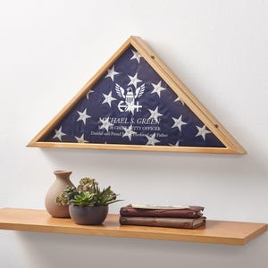 May include: A wooden triangular flag display case with a blue and white flag inside. The flag has white stars and an emblem. Text on the flag reads "MICHAEL S. GREEN, SENIOR CHIEF PETTY OFFICER, Dutiful and Proud Sailor, Husband and Father."