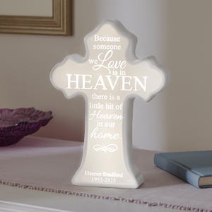 May include: White ceramic cross with a message that reads "Because someone we love is in Heaven, there is a little bit of Heaven in our home." The cross also has the name "Eleanor Bradford" and the dates "1952-2025" on the base.