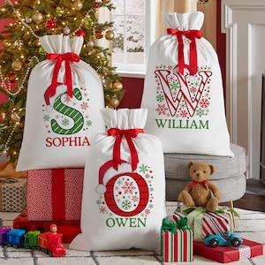 May include: Three white Christmas gift sacks with red ribbon ties. Each sack features a large initial and name in green and red, with snowflake accents. The sacks are filled and sit among wrapped gifts and a Christmas tree.