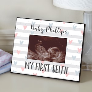 May include: A black picture frame with a white and gray striped background. The frame has a pink and blue heart pattern and the text "Baby Phillips My First Selfie".