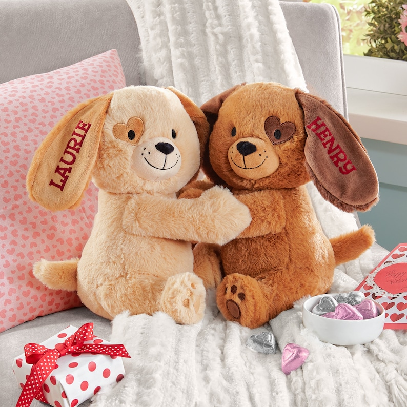 May include: Two plush brown and beige dog toys with embroidered hearts on their eyes. The dog on the left has the name "Laurie" embroidered on its ear and the dog on the right has the name "Henry" embroidered on its ear.