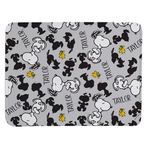 May include: A rectangular mat with a light gray background featuring a repeating pattern of Snoopy in white, black silhouettes of Snoopy, small yellow Woodstock figures, and the name Taylor printed in a black sans-serif font throughout the design.