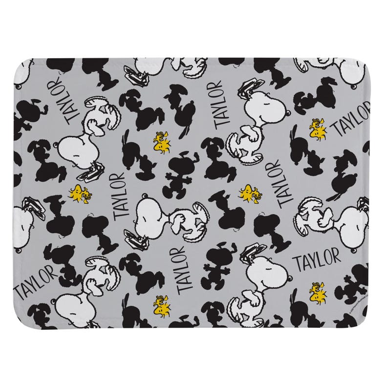 May include: A rectangular mat with a light gray background featuring a repeating pattern of Snoopy in white, black silhouettes of Snoopy, small yellow Woodstock figures, and the name Taylor printed in a black sans-serif font throughout the design.