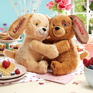 May include: Two plush brown and beige dog toys with embroidered hearts on their eyes and the names "Laurie" and "Henry" on their ears. The toys are hugging each other and are sitting on a table with a pink card that says "XOXO" and a plate of waffles.