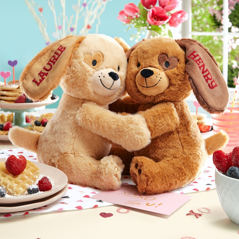 May include: Two plush brown and beige dog toys with embroidered hearts on their eyes and the names "Laurie" and "Henry" on their ears. The toys are hugging each other and are sitting on a table with a pink card that says "XOXO" and a plate of waffles.