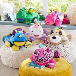 May include: A collection of colorful plush toys with embroidered names, including a green dragon, a pink hippo, a purple unicorn, a blue dinosaur, a brown dog, and a pink leopard. The toys are arranged on a white and yellow beanbag chair and a yellow ottoman.