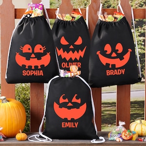 May include: Four black drawstring bags with orange pumpkin faces and names "Sophia", "Oliver", "Brady", and "Emily" printed on them. The bags are filled with candy and are hanging on a wooden fence.
