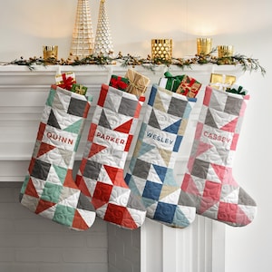 May include: Four quilted Christmas stockings featuring geometric triangle patterns in various color palettes. Each stocking is personalized with the names QUINN, PARKER, WESLEY, and CASSEY in block lettering, with small gift boxes tucked inside the top.