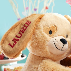 May include: A plush tan dog with a heart-shaped patch over one eye. The dog's ear is embroidered with the name "LAURIE".