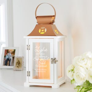 May include: White wooden lantern with a copper-colored top and handle. The lantern has a glass front with the text "A Light remains where a Beautiful Soul has passed". The lantern is lit from within, creating a warm glow.