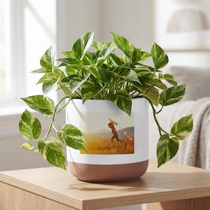 May include: A white and terracotta-colored ceramic planter pot with a printed image of a child running in a field. The pot is filled with a green and white variegated pothos plant. The pot sits on a wooden surface.