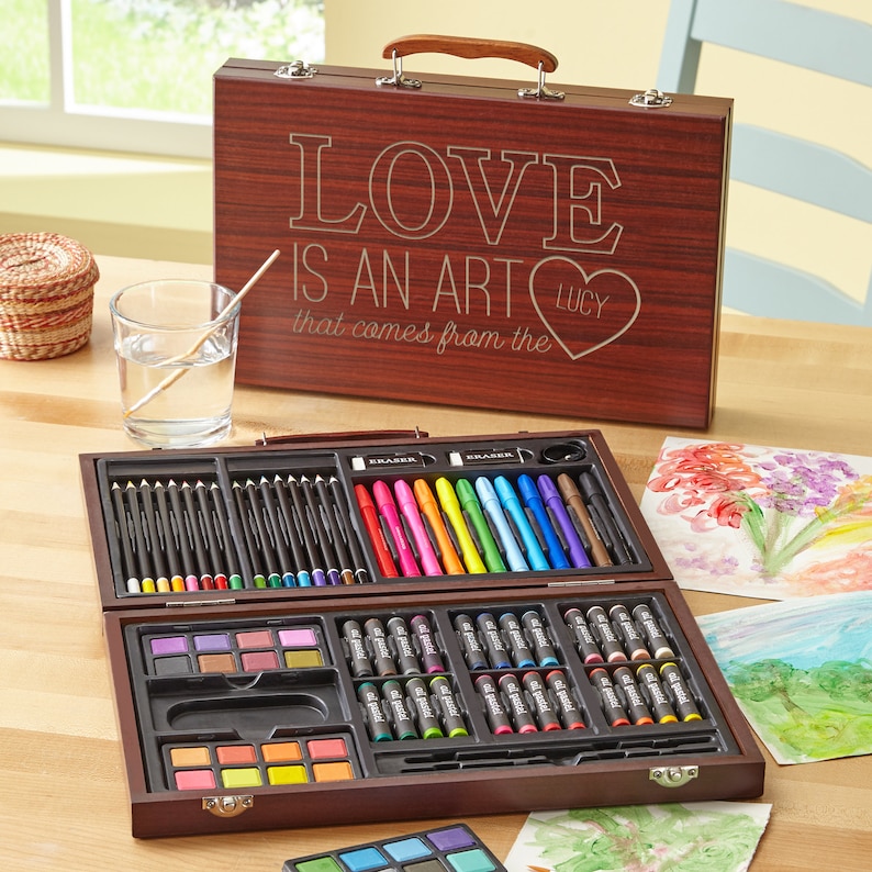 May include: A wooden art set with a brown carrying case. The case has the words "LOVE IS AN ART that comes from the" engraved on it, with a heart that says "LUCY". The set includes colored pencils, markers, crayons, and watercolor paints.