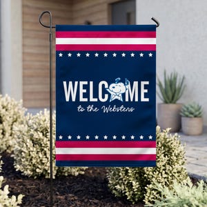 May include: A rectangular garden flag featuring a dark blue background with horizontal red and white stripes at the top and bottom. The flag displays white stars, the text WELCOME, a Snoopy graphic, and the text to the Websters in white.