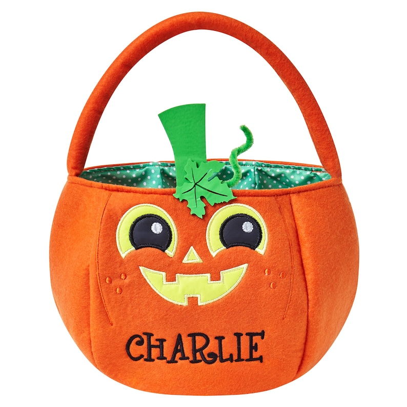 May include: Orange felt pumpkin-shaped Halloween treat bag with a green handle. The bag features a smiling jack-o'-lantern face with black eyes, yellow accents, and the name "CHARLIE" embroidered in black. The interior lining is green with white polka dots.