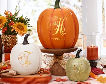 May include: Three decorative pumpkins featuring gold script lettering and botanical accents. A large orange pumpkin displays the letter H and the word HAPPINESS, a white pumpkin shows the letter F and FAMILY, and a gold glitter pumpkin shows the letter L.