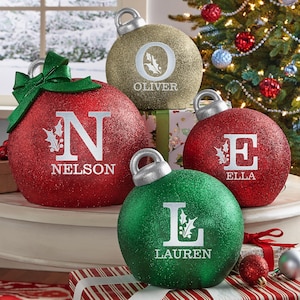 May include: Four large, glittery Christmas ornaments in red, gold, and green. Each ornament has a silver cap and is personalized with a name and initial. The red ornament on the left has a green bow. A Christmas tree is visible in the background.